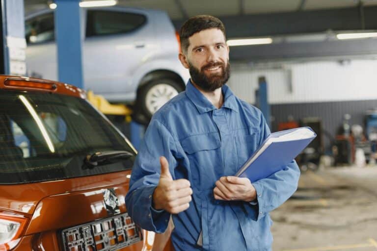 Auto mechanic in blue coveralls with clipboard giving thumbs up in garage.